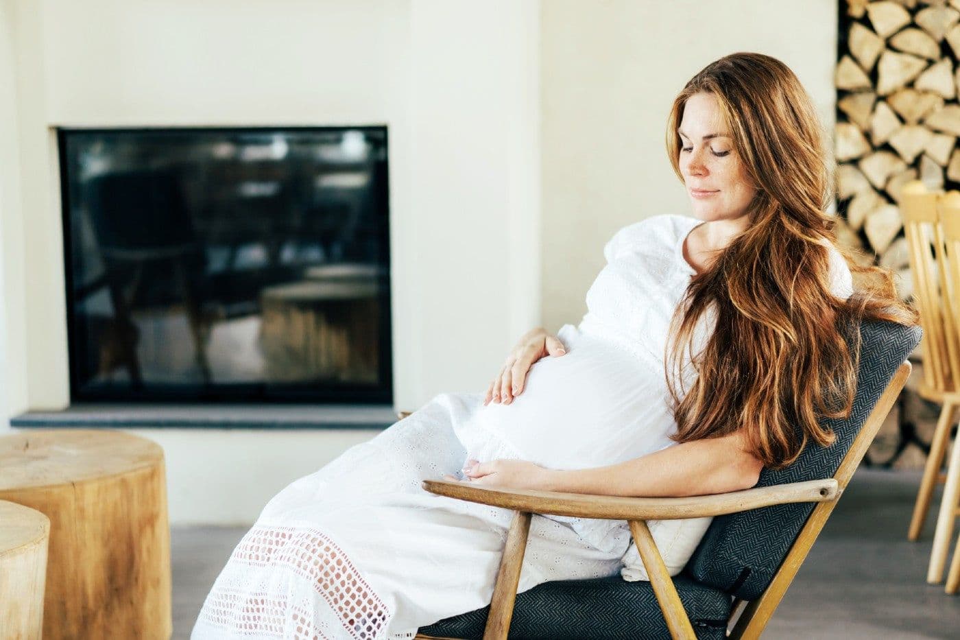 A pregnant woman sits by a fireplace while gazing at her belly