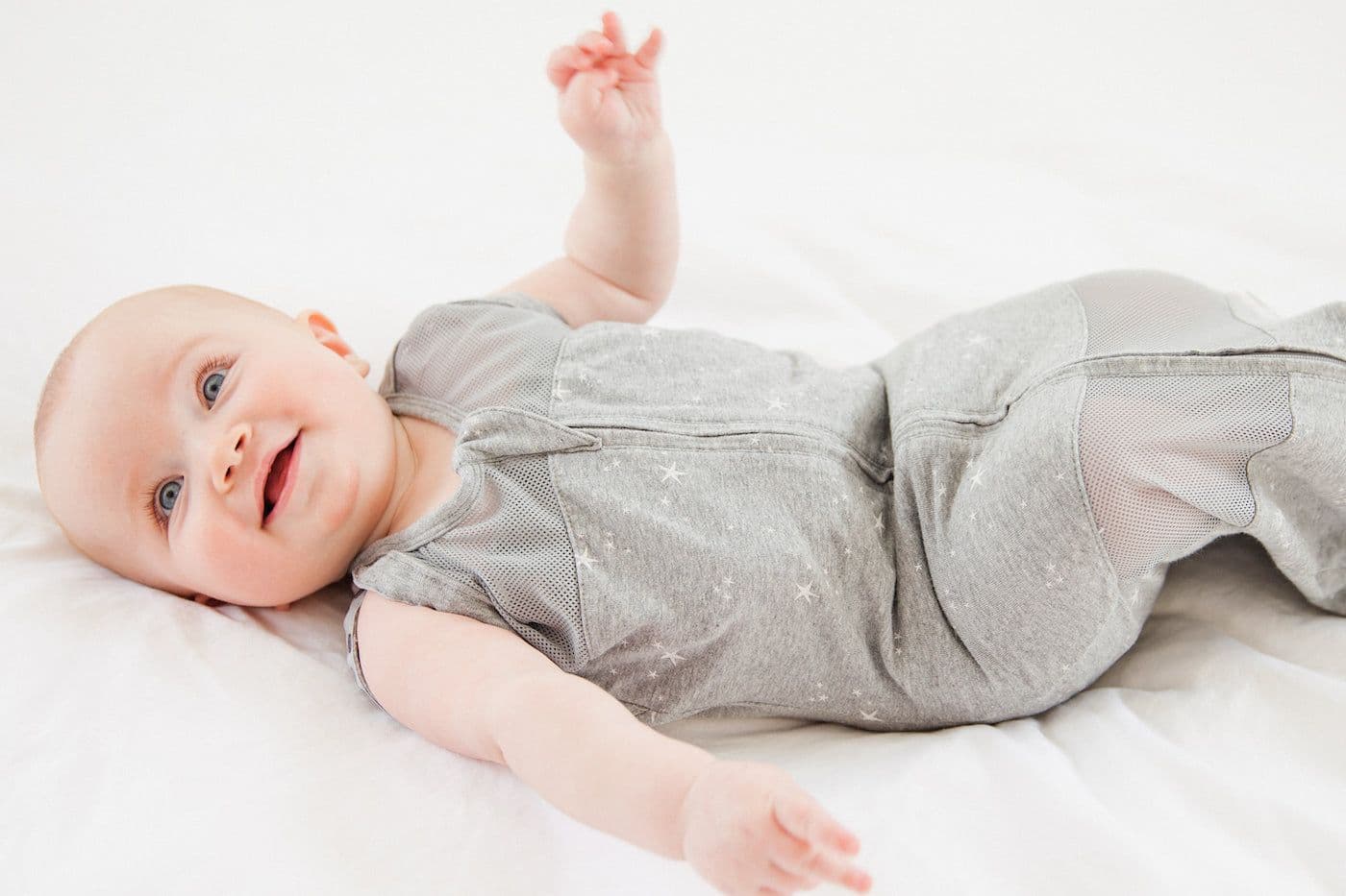 A smiling baby wears a grey Sleepea swaddle