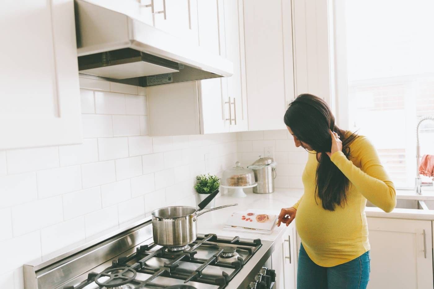 A pregnant woman looks at a cookbook as she prepares a meal on the stove.